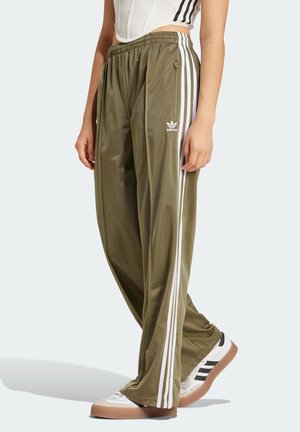 Femme portant un pantalon de survêtement Adidas vert olive avec des bandes blanches et des baskets blanches avec des bandes noires sur un fond clair.