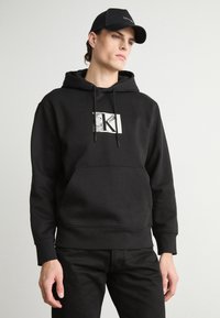 Black hooded sweatshirt with a front pocket, featuring a silver "CK" logo on a white rectangle. Paired with black jeans, showing a simple design.