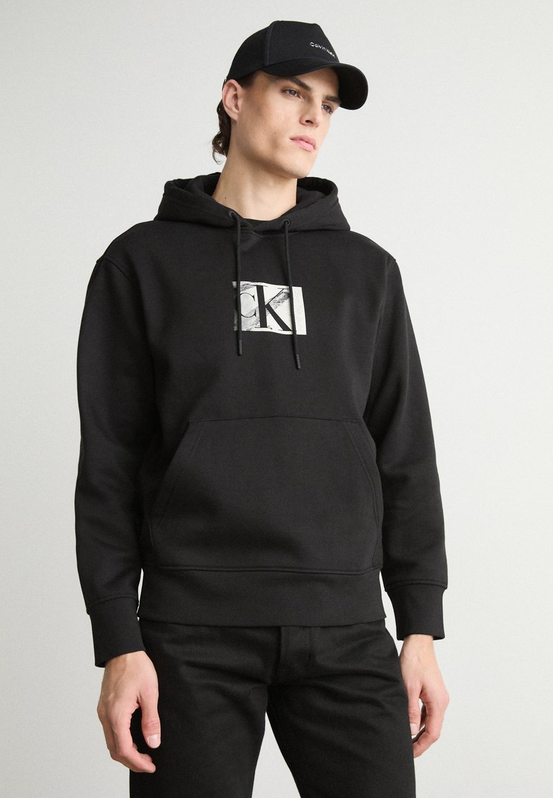 Black hooded sweatshirt with a front pocket, featuring a silver "CK" logo on a white rectangle. Paired with black jeans, showing a simple design.
