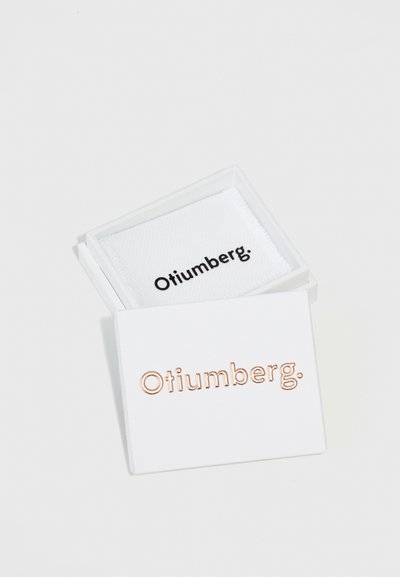 Hvid boks med et gennemsigtigt låg, der indeholder et stykke hvid stof. "Otiumberg." er prentet i rosaguld på låget. Teksten er klar og centreret.