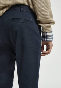 Pantalon marine en coton avec une texture lisse, doté d'une poche arrière avec bouton et d'une taille élastique, associé à un pull en tricot beige.