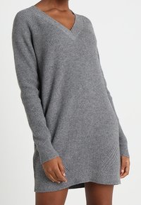 Robe pull grise en maille avec col en V, manches longues, texture côtelée et accents latéraux. Coupe décontractée et confortable, longueur au genou.