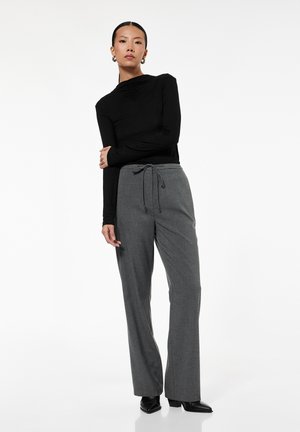 Kvinde stående med armene korsede iført sort langærmet top, grå bukser med snor og sorte spidse sko på hvid baggrund.