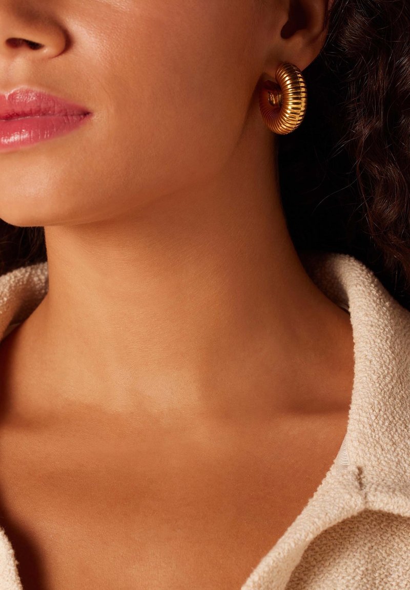 Gouden hoepeloorbellen met een ribbelstructuur en een cirkelontwerp. Een zachte huidtint en zachte stof van een beige top zijn zichtbaar.