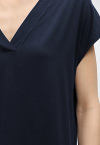 Κοντινό πλάνο ενός ατόμου που φοράει ένα κοντομάνικο μπλουζάκι σκούρου ναυτικού μπλε με V λαιμόκοψη, που δείχνει μέρος του μπράτσου και του άνω στήθους.