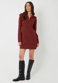 Vrouw in een lange mouw, roestkleurige trui-jurk met een ritskraag en zwarte leren laarzen tot de knie, staande tegen een witte achtergrond.
