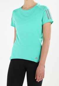 Maglietta sportiva verde menta a maniche corte con scollo rotondo, caratterizzata da strisce grigie sulle spalle e un emblema sull'orlo inferiore.