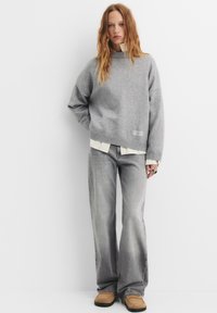 Maglione grigio oversize con collo alto, indossato sopra una camicia bianca. Abbinato a jeans grigi sbiaditi a zampa d'elefante e scarpe slip-on marrone chiaro.