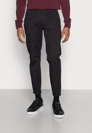 Uomo che indossa pantaloni neri slim fit, sneakers nere con suole bianche e una camicia a maniche lunghe rosso scuro, in piedi su un pavimento grigio.