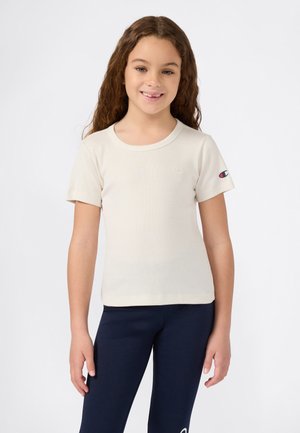 Fille souriante avec de longs cheveux bouclés portant un t-shirt blanc à manches courtes et un legging bleu marine sur un fond uni.