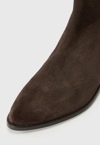 Botte cheville en suède brun avec un bout pointu, texture lisse et silhouette élégante. Présente une semelle en cuir noir fin et des détails de couture.