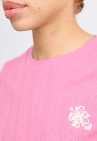 Rózsaszín pamut póló, textúrált mintával, bordás nyakkal és bal mellen fehér hímzett hibiszkusz virág részlettel.