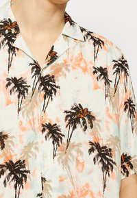 Camicetta a maniche corte con bottoni in tessuto leggero, caratterizzata da un motivo di palme tropicali in nero e arancione su uno sfondo pastello.