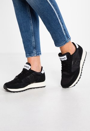 Person wearing black and white Saucony sneakers and blue jeans standing on a white floor with legs crossed.