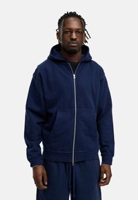 Dark navy zip-up hoodie in soft fabric with a kangaroo pocket, ribbed cuffs, and drawstring at the hem. Worn over a black shirt.