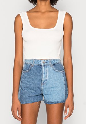 Kobieta w białym bezrękawnym krótkim topie i wysokich dżinsowych szortach z dwukolorowymi niebieskimi panelami, stojąca na jednolitym tle.