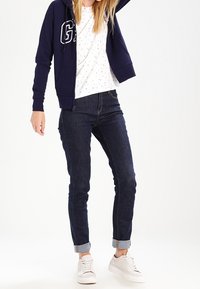 Sudadera con cremallera navy y logo, camiseta blanca con puntos, jeans de mezclilla oscuros y deportivas claras. Puños de los jeans remangados, ajuste casual.