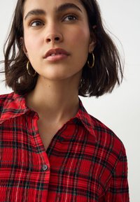 Rotes kariertes Hemd mit Knöpfen aus strukturiertem Stoff, mit Kragen, schwarzen Knöpfen und dezenten goldenen Fäden, die ins Muster eingewoben sind.