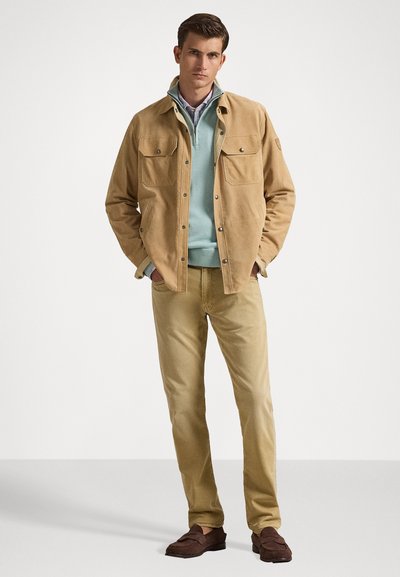 Καφέ σουέτ μπουφάν με δύο τσέπες στο στήθος, φορεμένο πάνω από ένα ανοιχτό μπλε πουλόβερ και πουκάμισο με γιακά, συνδυασμένο με μπεζ παντελόνι και καφέ loafers.