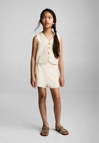 Gilet beige senza maniche con bottoni e ricami floreali, abbinato a pantaloni corti coordinati con bordi scallop. Indossato con sandali semplici.
