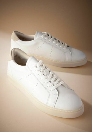 Tenisi din piele albă cu o tabără de călcâie din suede bej texturat. Au un design cu șireturi, perforații minime și o talpă groasă, translucidă.