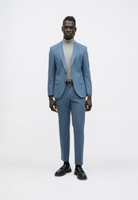 Világoskék öltöny, szabott blézerrel és testre szabott nadrággal, szürke garbóval párosítva. Fekete alkalmi cipők teszik teljessé a megjelenést.