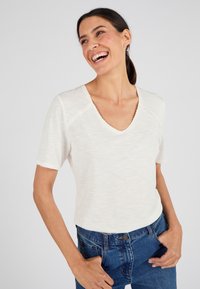 Camiseta blanca de manga corta con escote en V y borde festoneado, hecha de una tela texturizada, combinada con jeans de mezclilla azul.