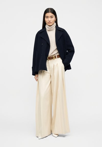 Γυναίκα που φοράει ναυτικό παλτό, μπεζ πουλόβερ ζιβάγκο, πλατιά κρεμ πλισέ παντελόνια, καφέ ζώνη και λευκά μυτερά παπούτσια, στέκεται με το ένα χέρι στην τσέπη.