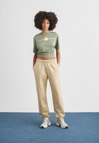 Ελιάς πράσινο crop t-shirt με λευκό λογότυπο, μπεζ φόρμα με ελαστική μέση και ανοιχτόχρωμα αθλητικά παπούτσια με υφή λεπτομέρειες.