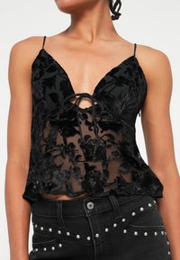 Melna ziedu mežģīņu blūze ar caurspīdīgu audumu, dziļu V veida kakla izgriezumu un regulējamiem spaghetti siksniņām. Priekšā mezglota detaļa, saskaņota ar augsto jostasvietu melnām džinsām.