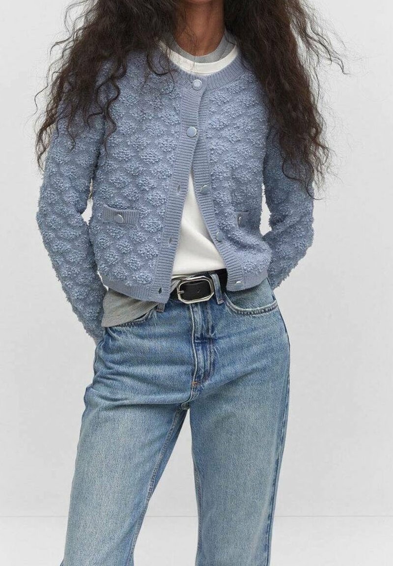 Personne portant un cardigan boutonné texturé bleu clair sur des hauts superposés blancs et gris, rentrés dans un jean bleu taille haute avec une ceinture noire.