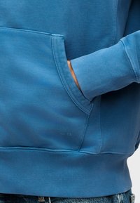 Το χέρι ενός ατόμου είναι μερικώς μέσα στην μπροστινή τσέπη ενός μπλε φούτερ με ριμπ μανσέτες και τελείωμα, συνδυασμένο με τζιν παντελόνι.