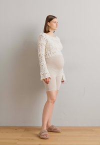 Krošu adīts krēmsvārds ar plašām piedurknēm, saskaņots ar bežām velosipēda šortiem un pūkainām bežām čībām. Modelis stāv pret gaismas sienu.
