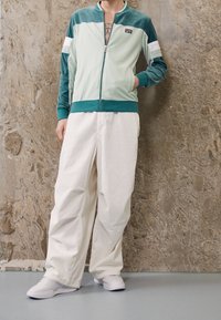 Jasnozielona kurtka zapinana na zamek z ciemnoturkusowymi akcentami i białym paskiem, zestawiona z luźnymi, kremowymi spodniami i białymi trampkami na gładkiej powierzchni.