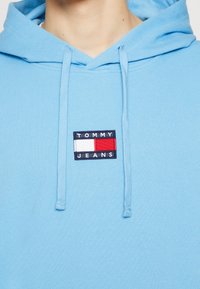 Ljusblå hoodie med dragsnören och en framficka. Har en rektangulär logotyp i marinblått, rött och vitt med texten "TOMMY JEANS."