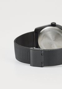 Reloj con correa de malla negra, con una caja negra hexagonal y un fondo de acero inoxidable, que presenta un grabado de "Hugo" y un cierre deslizante.