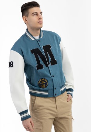 Giacca varsity blu e bianca con un grande emblema nero "M", dotata di una toppa sul lato e polsini e orlo a righe a coste.