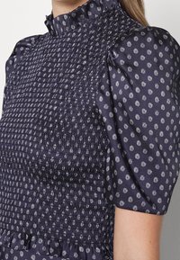 Robe bleu marine avec des motifs circulaires blancs, corsage froncé, col montant à volants et manches courtes bouffantes portée par une personne.