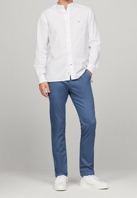 Weißes Hemd mit Kragen, kombiniert mit hellblauen Hosen. Beide Kleidungsstücke haben eine glatte Textur; die Schuhe sind weiße Sneakers.