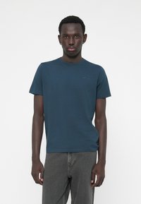 Tricou din bumbac de culoare bleumarin, cu guler rotund, mâneci scurte și un detaliu discret al logo-ului pe piept, purtat cu pantaloni din denim gri.