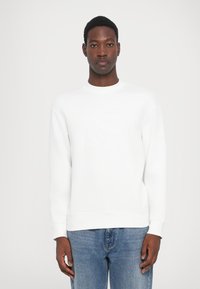 Sudadera blanca con puños y dobladillo de canalé, con un detalle sutil de logo en relieve en la parte frontal. Combinada con jeans de un azul claro.