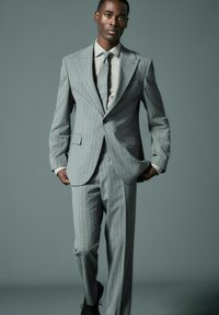 Traje gris claro con rayas, con chaqueta de un solo botón y pantalones de frente plano, combinado con una camisa de color claro y una corbata texturizada.