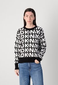 Juodas ir baltas raštuotas megztinis su pasikartojančiu "DKNY" logotipu; banguoti rankogaliai ir apačia; ilgomis rankovėmis; dėvimas su šviesiai mėlynomis džinsomis.