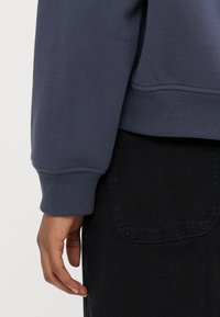 Polsino di felpa blu navy con trama a coste, abbinato a un capo nero con tasca. Dettaglio sulla manica e sul materiale.