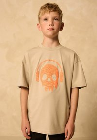Béžové tričko s krátkym rukávom s popraskanou oranžovou lebkou a slúchadlami na prednej strane. Tkanina vyzerá mäkko a má uvoľnený dizajn.