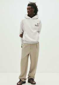 Weißer Kapuzenpullover mit Fronttasche, auf dem das Logo "Mickey's" abgebildet ist. Kombiniert mit hellbeigen, weit geschnittenen Hosen und dunklen Sneakers. Glatte Textur.