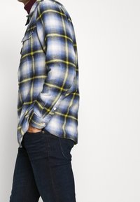 Uomo che indossa una camicia a quadri blu, bianca e gialla con le mani nelle tasche dei jeans scuri, in piedi davanti a uno sfondo bianco uniforme.