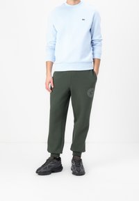 Sweat-shirt bleu clair avec encolure ronde et poignets côtelés, associé à un pantalon de survêtement vert foncé avec un logo. Des baskets noires texturées complètent le look.