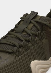 Κοντινό πλάνο αθλητικού παπουτσιού σε ελιά πράσινο χρώμα με υφασμάτινο διχτυωτό υλικό, χοντρές κορδόνες και λεπτομέρεια στη σόλα σε μπεζ χρώμα με ανάγλυφο σχέδιο.