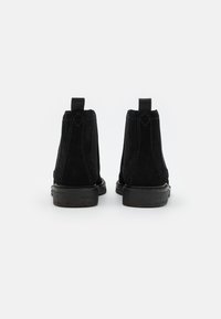 Schwarze Wildleder-Ankelboots mit runder Zehenpartie, flacher schwarzer Gummisohle und Zugschlaufe an der Rückseite. Vertikale Steppnähte an den Seiten.